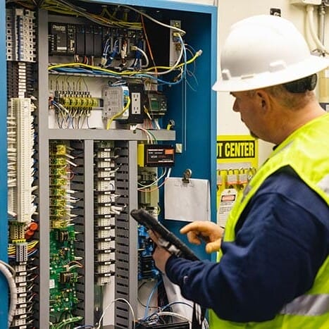 a man in a hardhat and neon vest looking at a utility control center with wires and buttons