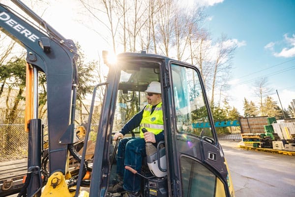 Man controlling a backhoe in a parking lot with trees behind