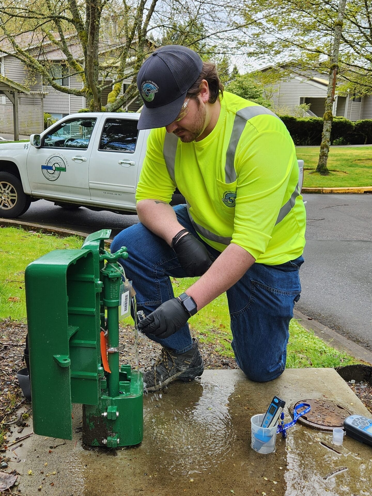 A worker in a neon yellow shirt and gloves kneels by a green utility box on a sidewalk, preparing a water testing sample. A white truck is parked nearby.